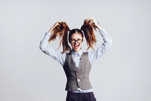 Una divertida colegiala joven con gafas y un chaleco gris juega con su pelo, se ve feliz — Foto de Stock