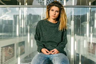 Kameraya bakarak kalın sarı saçlı bir cam çitte oturan şık genç kız