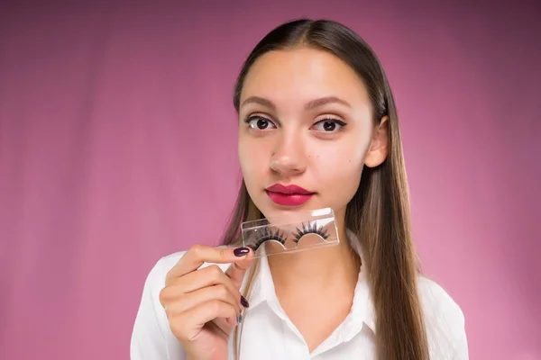 Belle jeune fille dans une chemise blanche, avec du rouge à lèvres rose sur ses lèvres tenant dans sa main un paquet de faux cils — Photo