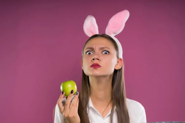 La chica sorprendida en las orejas del conejo mira hacia adelante con una manzana en sus manos — Foto de Stock