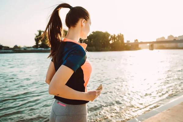 Unga långhåriga fitness tjej i sportkläder körs vid floden, i solen, lyssna på musik i hörlurar — Stockfoto