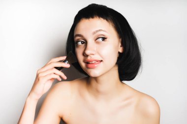 Kısa siyah saç ve temiz cilt ile çekici bir genç kız bir fırçayla yüzünde makyaj uygulanır