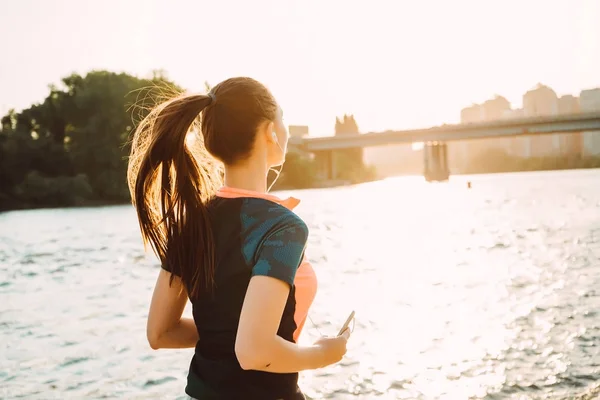 Långhåriga unga fitness tjej körs vid floden, i solnedgången, lyssna på musik i hörlurar — Stockfoto