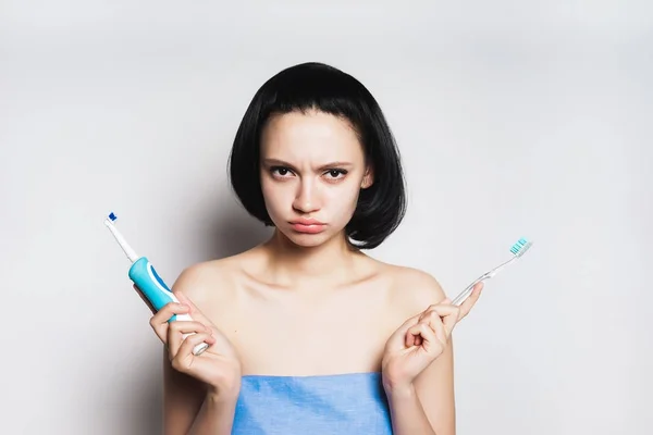 a sad young girl with black hair is holding an electric toothbrush and the usual one, and she can not choose