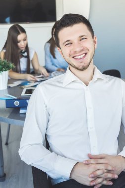 gülüyor, onun arkasında Beyaz gömlekli iki kız bir masada çalışma başarılı genç erkek ofis çalışanı