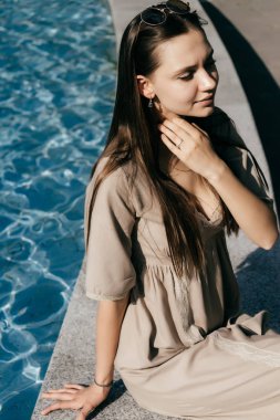 uzun saçlı çekici model kız güneş sahiptir, mavi su ile havuz başında oturur