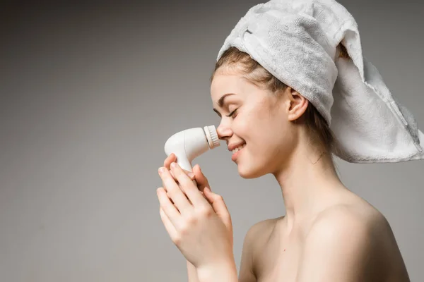 Divertente ragazza con un asciugamano bianco sulla testa pulisce la pelle sul suo viso con una spazzola elettrica — Foto Stock