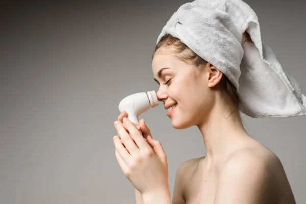 Funny young girl with a white towel on her head cleans the skin on her face with an electric brush — Stock Photo, Image