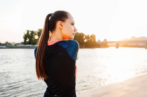 Jovem menina fitness de cabelos longos amassando músculos, pelo rio ao pôr do sol — Fotografia de Stock