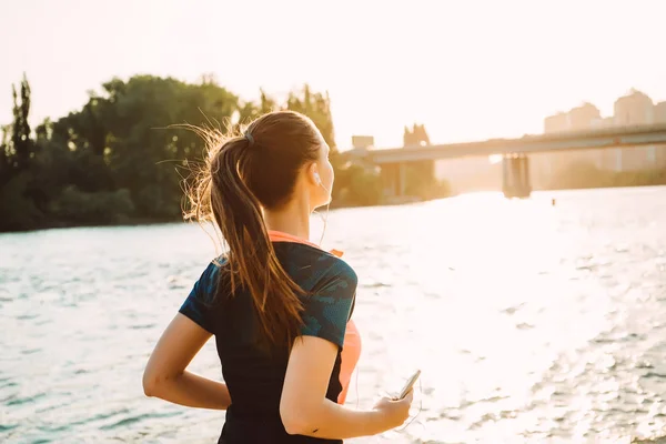 Smal tjej i sportkläder löper runt floden, lyssna på musik i hörlurar — Stockfoto