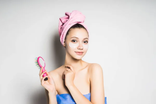 Νεαρή κοπέλα φρέσκια με μια ροζ πετσέτα στο κεφάλι της, κάτω από τα μάτια των patches σιλικόνης, κατέχει μια χτένα — Φωτογραφία Αρχείου