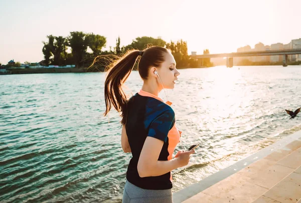 Sport flicka med telefonen i händerna körs vid solnedgången — Stockfoto