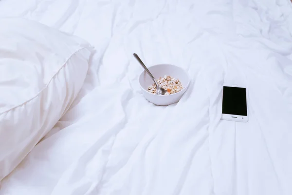 Na łóżku arkusz biały tam jest płyta płatków owsianych na śniadanie, leży smartfon — Zdjęcie stockowe