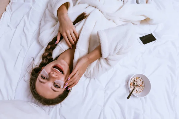 Gadis muda yang bahagia bangun, berbaring di tempat tidur, di bawah mata patch silikon, sarapan di tempat tidur — Stok Foto