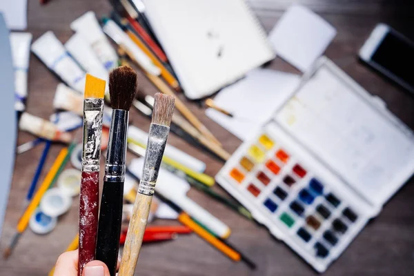 Sobre a mesa estão espalhados materiais de arte, tintas aquarelas coloridas, lápis, guache, tubos com tinta a óleo, a menina segura um pincel na mão — Fotografia de Stock