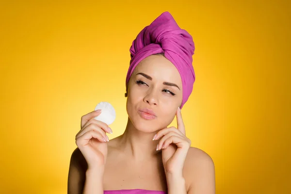A young girl with a pink towel on her head cleans the skin on her face with a cotton pad, thinks about facial care — Stock Photo, Image