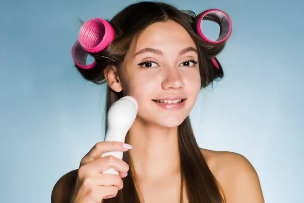 Ένα νεαρό όμορφο κορίτσι πρόκειται να ένα κόμμα, στο κεφάλι του ένα ψαλίδι για μπούκλες, καθαρίζει το πρόσωπό της με μια ηλεκτρική βούρτσα — Φωτογραφία Αρχείου