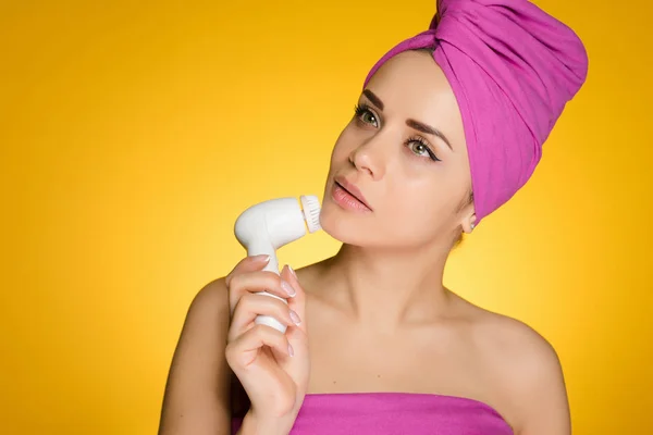 Μια νεαρή κοπέλα φαίνεται μετά το πρόσωπό της, καθαρίζει αυτό με μια ηλεκτρική βούρτσα, ένα ροζ πετσέτα στο κεφάλι της — Φωτογραφία Αρχείου