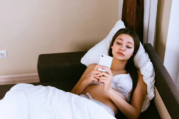 Una joven se acuesta en la cama y mira en su teléfono inteligente, se despierta temprano, disfruta del fin de semana — Foto de Stock