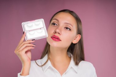 Genç kız pembe bir arka plan üzerinde sahte kirpik bir kümesini onun elinde tutar.