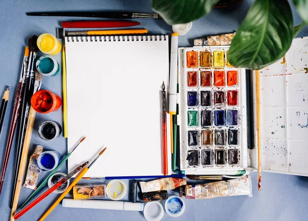 Em uma mesa azul colocar suprimentos de arte, um bloco de notas branco, aquarelas, guache e escovas — Fotografia de Stock