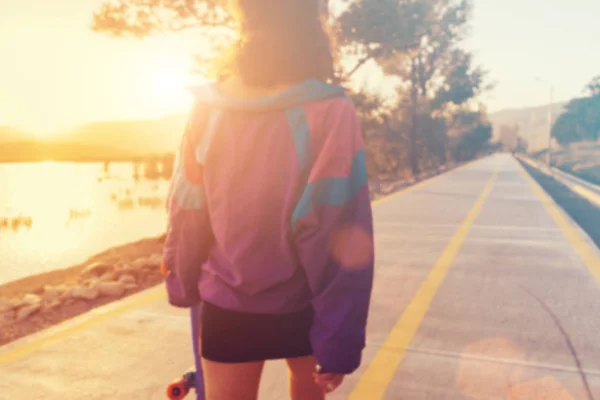 Stijlvolle mode meisje in een grote jas in de zon, in haar hand wandelingen paars is een skateboard, longboard — Stockfoto