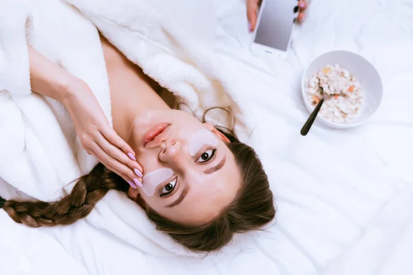 Uma jovem acordou cedo, deita-se na cama, sob os olhos de adesivos de silicone, há uma tarlet com aveia para o café da manhã — Fotografia de Stock
