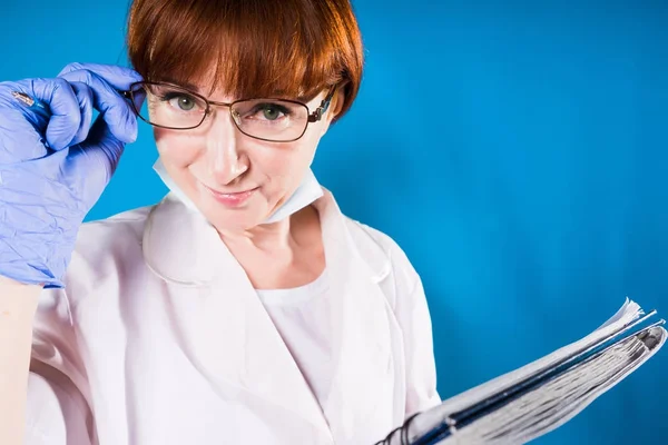 Una bella infermiera con gli occhiali tiene gli occhiali con la mano e guarda attentamente nella fotocamera e tiene una spessa cartella tra le mani, isolata su uno sfondo blu — Foto Stock