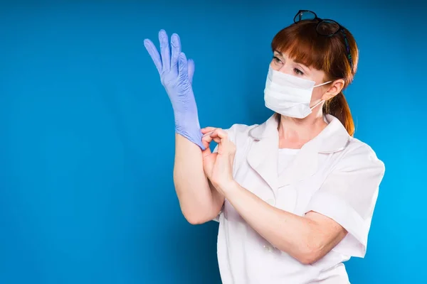 En kvinna i en medicinsk vit rock och bär en mask sätter på sina handskar och ser på händerna, isolerad på en blå bakgrund — Stockfoto