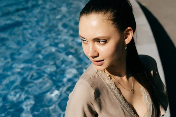 Hermosa chica se sienta junto a la piscina y se ve lindo a un lado. clima cálido de verano, día soleado — Foto de Stock