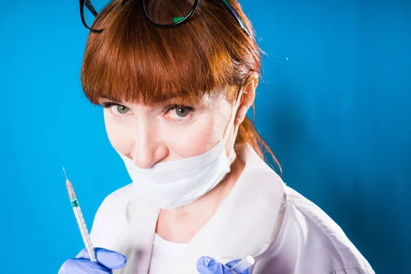 Ragazza dai capelli rossi un'infermiera in maschera tiene una siringa tra le mani e astutamente guarda nella fotocamera, isolata su uno sfondo blu — Foto Stock