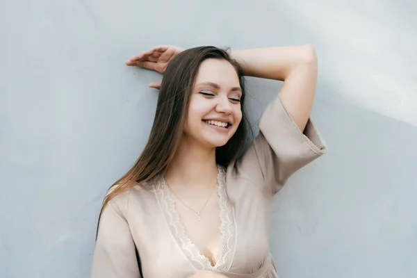 Linda chica en un vestido beige con el pelo largo de pie junto a la pared y riendo con los ojos cerrados — Foto de Stock