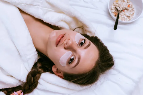Uma jovem acordou cedo, deitada em uma cama, sob os olhos de manchas de silicone, um prato de mingau de aveia café da manhã — Fotografia de Stock