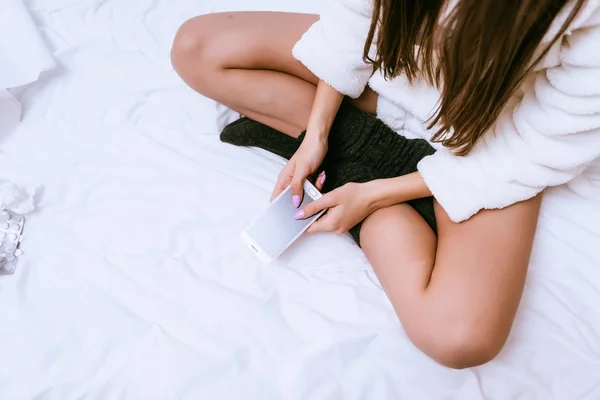 Девушка в белом махровом халате и черных теплых носках сидит на кровати, держа в руках смартфон — стоковое фото