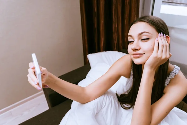 素敵な若い女の子に早起き、毛布の下のベッドに座っているし、selfie になります — ストック写真