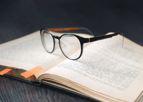 Σε μια σκούρα ξύλινη επιφάνεια βρίσκεται ένα βιβλίο με τους σελιδοδείκτες και τα γυαλιά σε μαύρο rim — Φωτογραφία Αρχείου