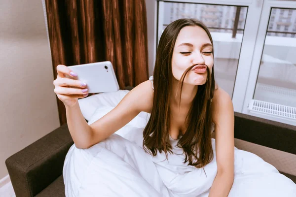 Gadis muda lucu duduk di tempat tidur di bawah selimut, membuat selfie — Stok Foto