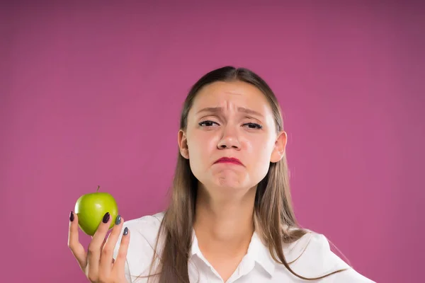 Aranyos lány úgy néz ki, a kamera nemtetszését, és apple tart a kezében, elszigetelt rózsaszín háttér — Stock Fotó