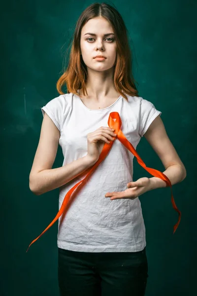 Menina séria em uma camiseta branca segura uma fita laranja na forma de um laço perto do coração e olha para a câmera — Fotografia de Stock