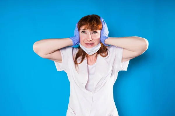 Eine wütende Krankenschwester mit Maske und Handschuhen schließt die Ohren mit den Händen und blickt isoliert auf blauem Hintergrund in die Kamera — Stockfoto