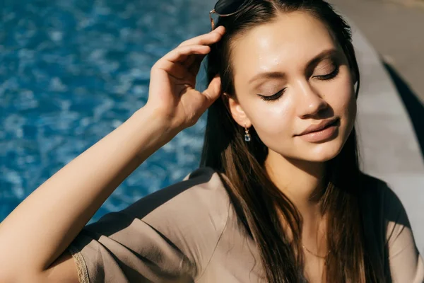 Una ragazza carina con un vestito beige si siede a bordo piscina e gode di una calda giornata estiva con gli occhi chiusi — Foto Stock