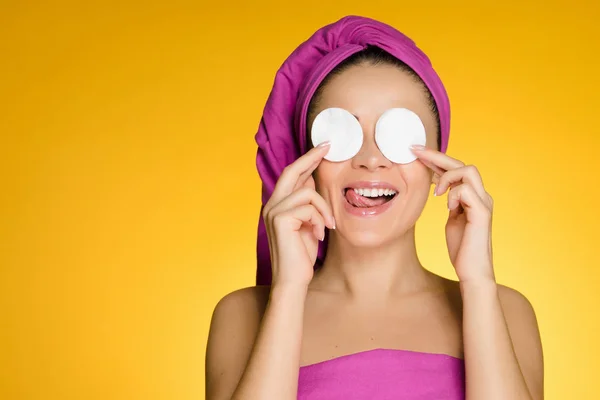 Heureuse jeune fille avec une serviette rose sur la tête nettoie la peau sur son visage avec des tampons de coton blanc sur un fond jaune — Photo