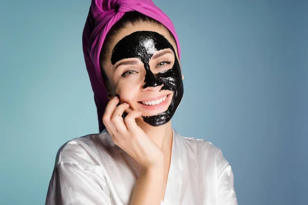 Happy νεαρό κορίτσι φροντίζει το δέρμα στο πρόσωπό της με μια μαύρη μάσκα, χαμογελώντας — Φωτογραφία Αρχείου