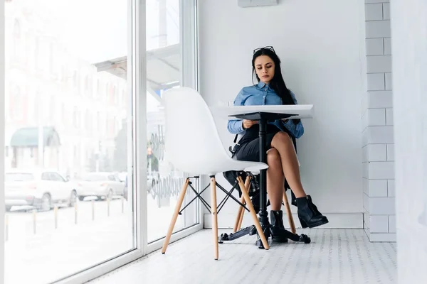 Успешная занятая девушка в синей рубашке и очках, фрилансер, сидит за столом в кафе — стоковое фото