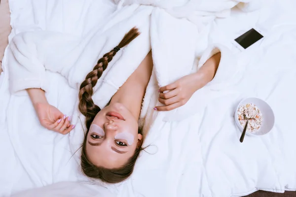 Una joven se despertó temprano, acostada en la cama, bajo los ojos de parches de silicona, junto a un plato con una útil avena —  Fotos de Stock