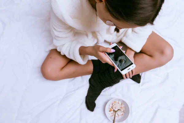 Młoda dziewczyna w szlafrok frotte biały jest posiadanie śniadanie w łóżku, fotografowanie jej płyta Kasza płatki owsiane — Zdjęcie stockowe