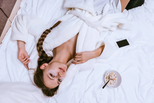 Une jeune fille en robe de chambre éponge blanche récemment réveillée, couchée au lit, sous les yeux de plaques de silicone, à côté d'une assiette avec un bouillie d'avoine utile — Photo