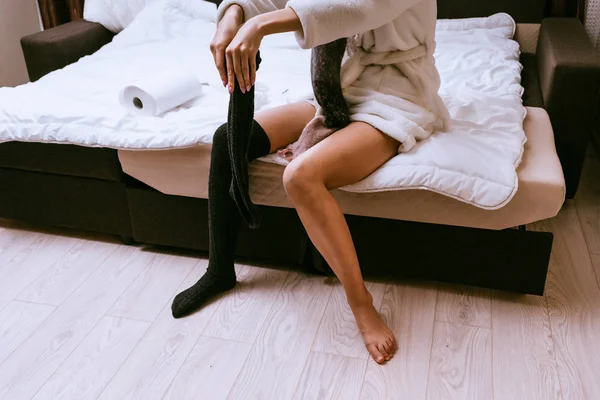 A young girl in a white terry dressing gown recently woke up, puts on warm black stockings on her feet — Stock Photo, Image