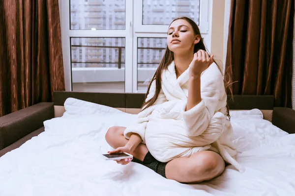 Девушка в белом махровом халате сидит на кровати, недавно проснувшись, держит в руке смартфон — стоковое фото