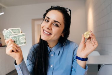 tutan gözlüklü başarılı genç iş kadını altın bitcoin ve dolar, gülümseyen ve kameraya bakarak Kripto para birimi hakkında düşünme